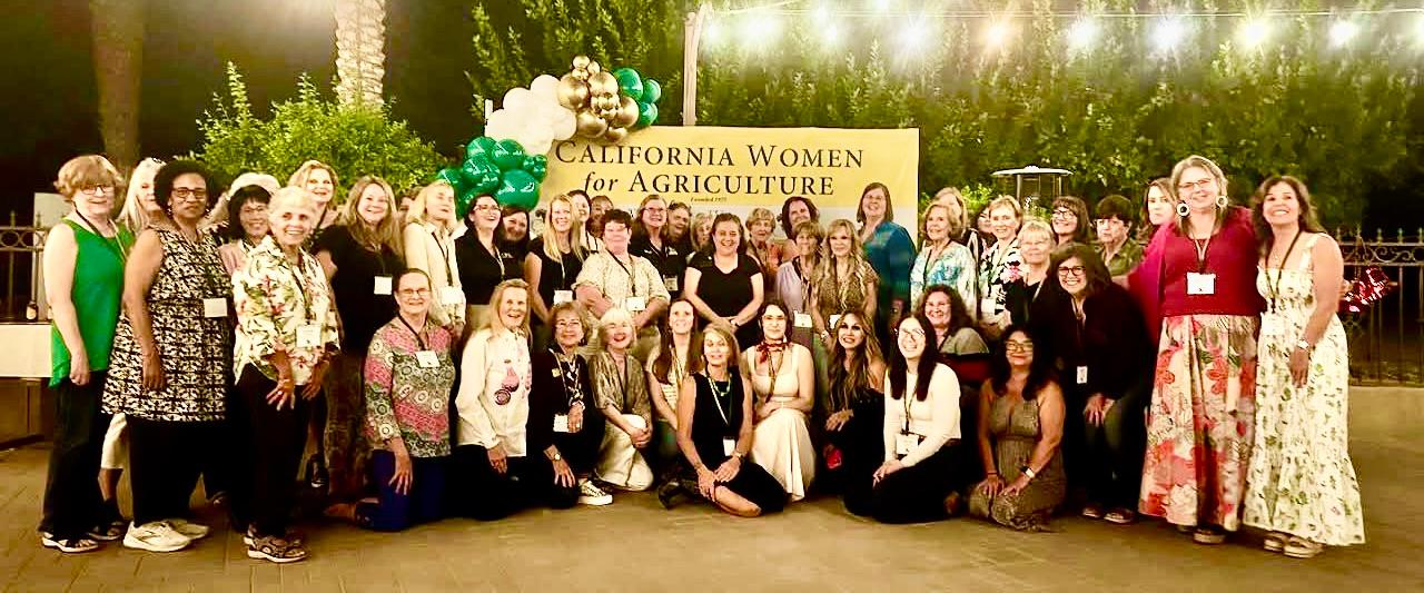 A large group of women pose together outdoors at night in front of a sign that reads “California Women for Agriculture.” The group is arranged in several rows, smiling at the camera, with some standing and others kneeling in front. Green and white balloons form an arch near the sign, and string lights hang overhead. Palm trees and greenery are visible in the background, creating a festive, celebratory atmosphere.